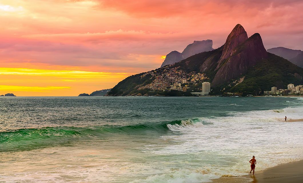 A vibrant sunset over Ipanema Beach in Rio de Janeiro, Brazil, with waves crashing on the shore, mountains in the background, and a few people walking along the sand—a perfect scene for a memorable Brazil tour.