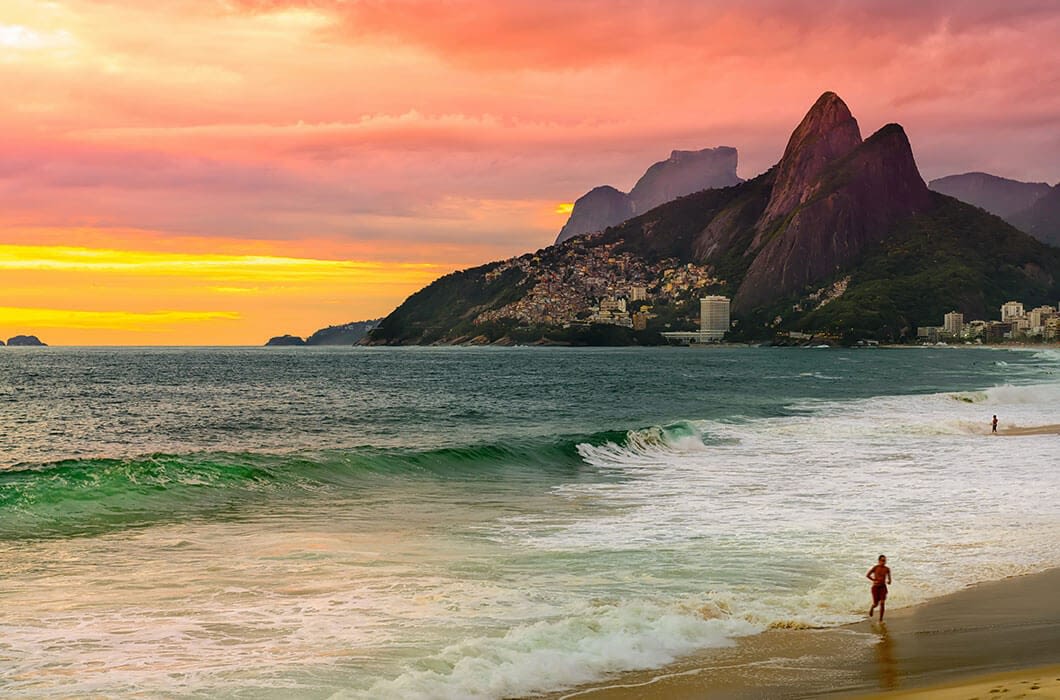 Colorful sunset over a beach with waves, a person walking along the shore, and tall mountains in the background near a coastal city—perfect for capturing memories on your Brazil Tour.