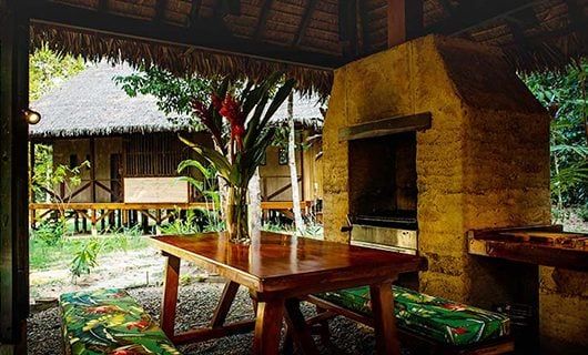 Outdoor dining area at Refugio Amazonas with a wooden table, benches with green floral cushions, tropical flowers, and a rustic clay oven under a thatched roof, surrounded by lush Amazon Rainforest greenery and a wooden building in the background.