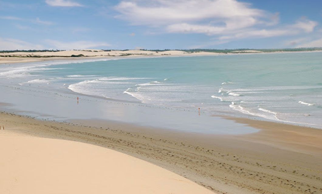 Expansive sandy beach with gentle waves rolling onto the shore under a blue sky with scattered clouds—perfect for a Luxury Adventure tour of Lencois Maranhenses, where rolling sand dunes and sparse vegetation frame the stunning landscape.