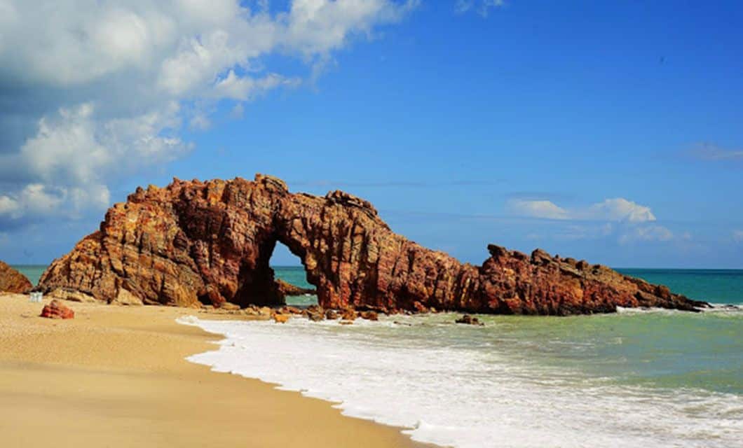 A natural rock formation with a large arch sits on a sandy beach, bordered by gentle ocean waves and backed by a blue sky with some clouds—perfect for a luxury adventure tour inspired by the wonders of Lencois Maranhenses.