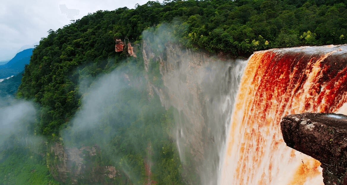 A wide, powerful waterfall with reddish-brown water cascades down a lush green cliffside, surrounded by mist and dense forest.