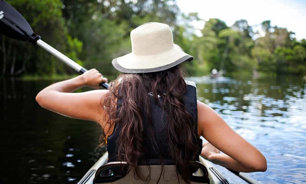 Person wearing a sunhat and life vest paddles a kayak on a calm river surrounded by lush green trees in Wild Argentina, viewed from behind.