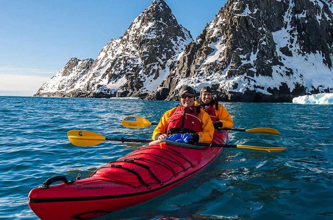 Two people in colorful jackets kayak in a bright red kayak on blue water, surrounded by snow-covered mountains and rocky cliffs under a clear sky—a spectacular family journey at the end of the world.
