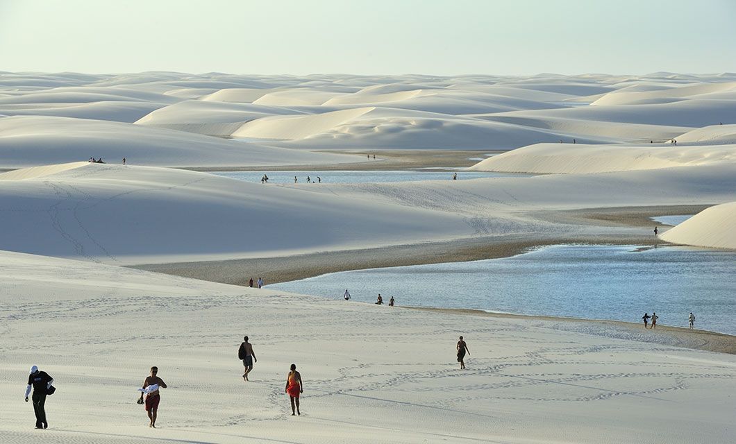People walk across the white sand dunes of Lencois Maranhenses, scattered with blue lagoons under a bright sky. Small groups and footprints dot this stunning landscape, perfect for a luxury adventure tour.