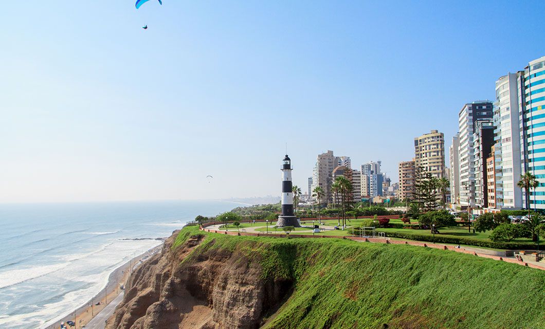 A black-and-white striped lighthouse stands on a green cliff overlooking the ocean, with modern high-rise buildings, palm trees, and a blue sky in the background—inviting travelers en route to Machu Picchu or a Galapagos Cruise to pause and take in the view.