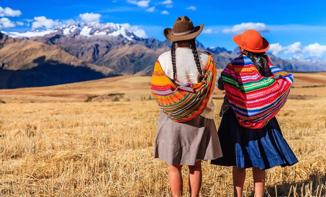 Two women in traditional Andean clothing and hats stand in a dry field, colorful shawls over their shoulders, gazing at the distant snow-capped mountains of the Sacred Valley beneath a bright blue sky with scattered clouds.
