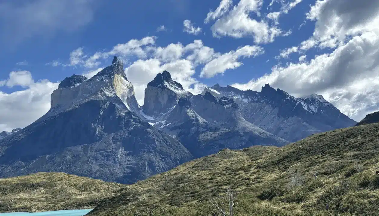 Dramatic mountain peaks with sharp ridges rise under a bright blue sky with scattered clouds, overlooking rocky green hills and a small turquoise lake—one of Patagonia’s natural wonders in South America.