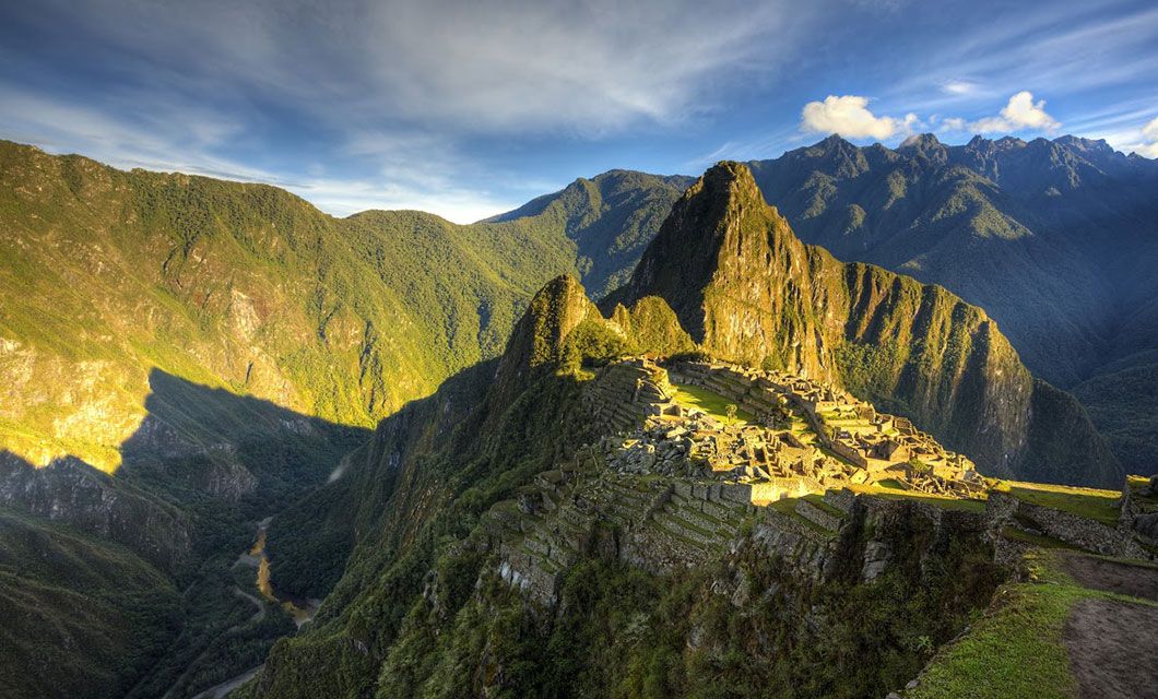 A panoramic view of Machu Picchu, one of South America's World Wonders, showcases the ancient Incan citadel with stone ruins, lush green terraces, and surrounding mountain peaks under a partly cloudy sky.