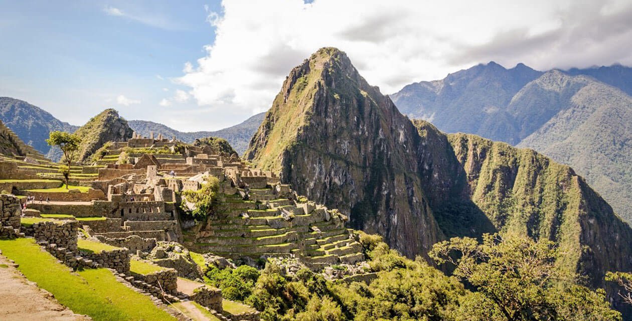 Ancient stone ruins of Machu Picchu sit on a green mountainside in South America under a partly cloudy sky, with the iconic Huayna Picchu peak rising dramatically—a breathtaking view for trekking enthusiasts.