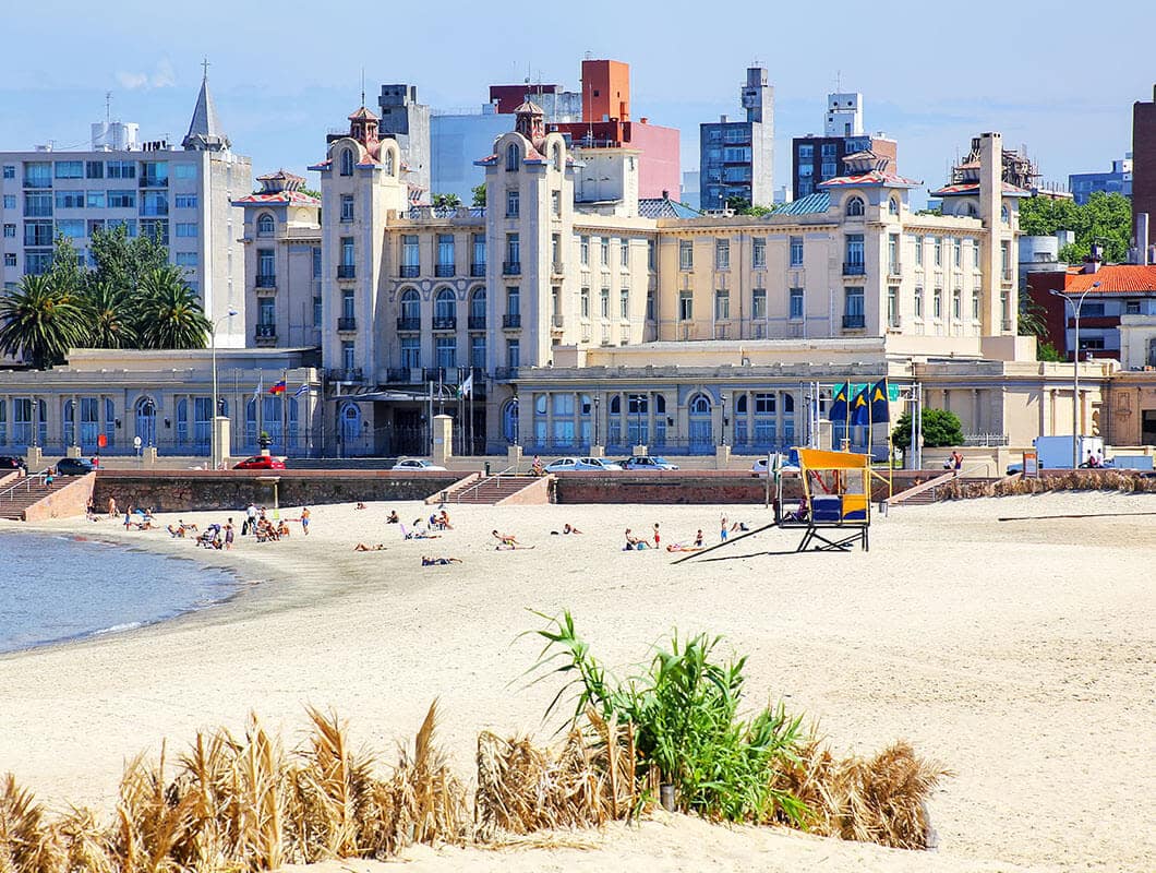 A sandy beach with people relaxing near the water. In the background, a large historic building with ornate architecture stands, reminiscent of those found in Buenos Aires, with city buildings behind and some greenery visible in the foreground.