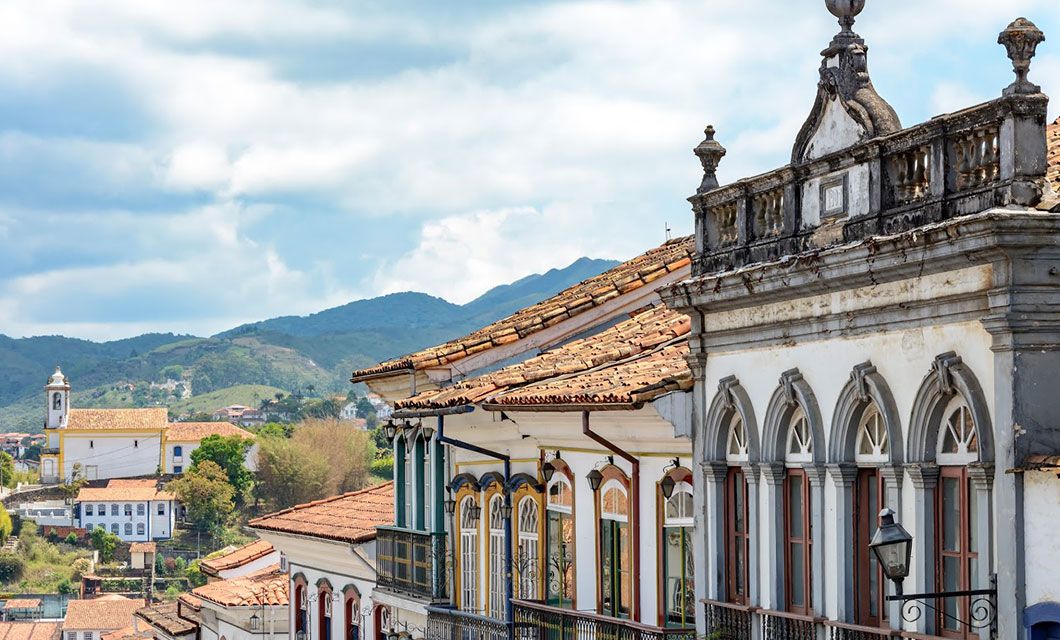 Colorful colonial buildings with arched windows line a street along the historic Route of Gold, with mountains and a church visible in the background under a partly cloudy sky.