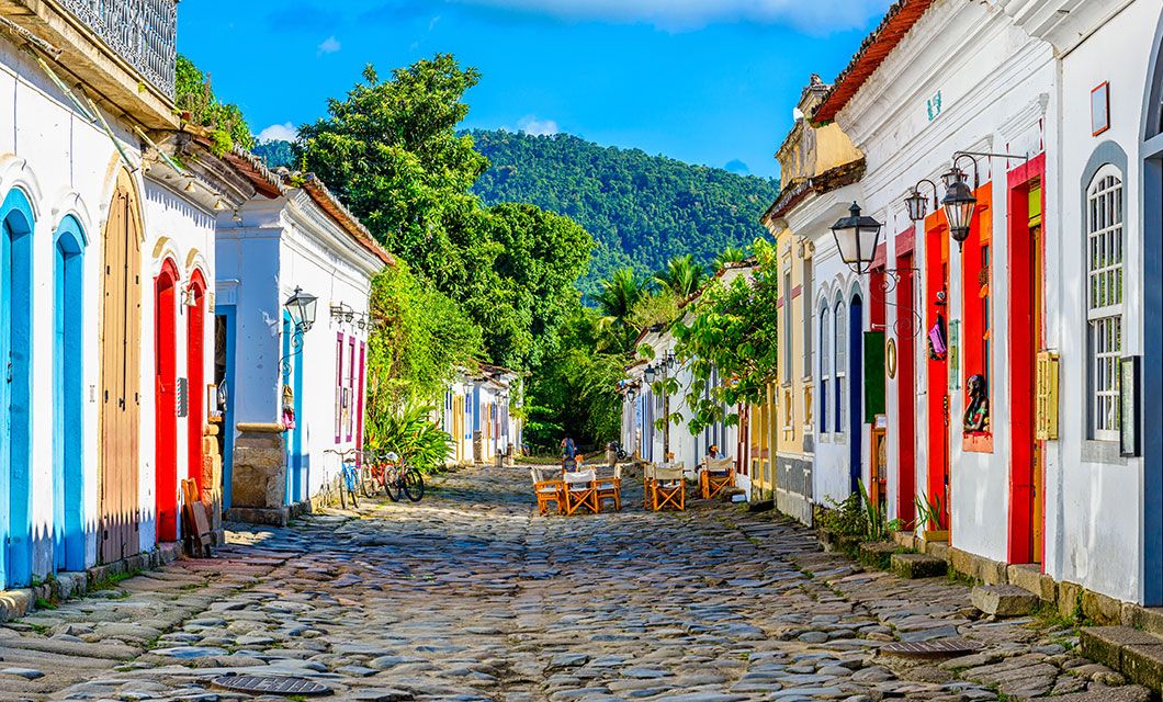 A cobblestone street along the historic Route of Gold, lined with colorful colonial-style buildings, vibrant doors and windows, outdoor tables, lush greenery, and hills in the background under a bright blue sky.