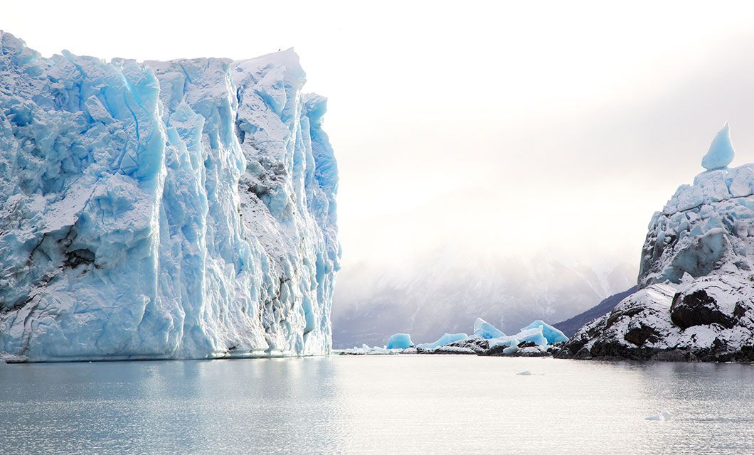 A large blue glacier rises from calm water near the Carretera Austral, with snow-covered rocks and smaller icebergs nearby under a misty, bright sky.