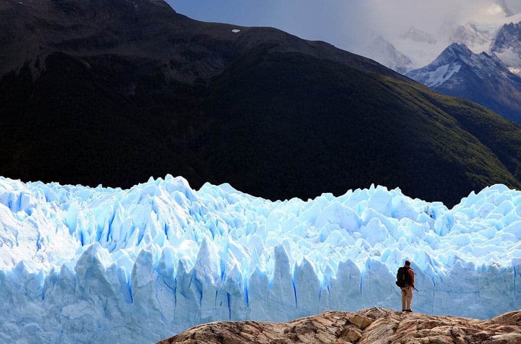 A True Explorer stands on rocky ground in Argentina, facing a massive, jagged blue glacier with forested mountains and snow-capped peaks in the background under a cloudy sky.