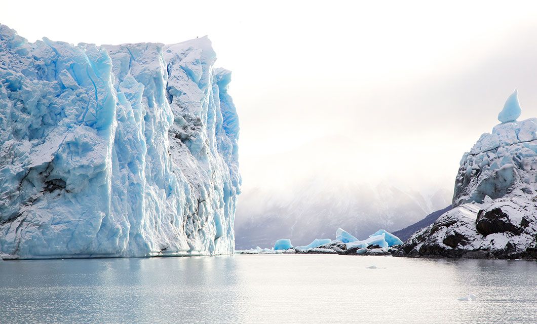 A large, blue glacier rises above calm water in Argentina, with jagged ice formations and snowy mountains in the background under a bright, cloudy sky—an iconic scene for any Argentina travel adventure.