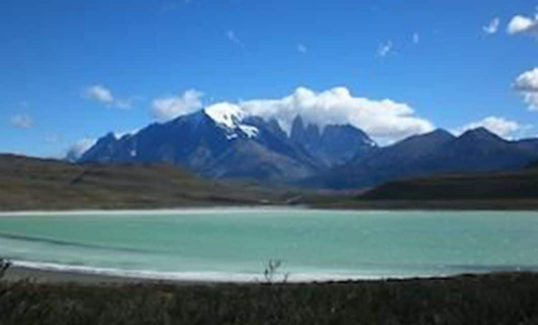 A turquoise lake with a mountainous backdrop under a bright blue sky, some peaks covered in snow and clouds, with rolling hills in the foreground—an iconic scene along the Carretera Austral on a classic Patagonia tour.