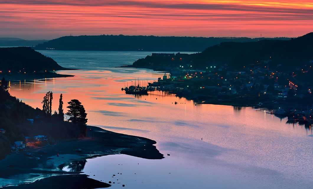 A tranquil river winds between hills in Northern Patagonia at sunset, reflecting vibrant orange and pink skies. Small lights from houses and boats dot the shoreline, creating a peaceful, scenic evening atmosphere for overland explorers.