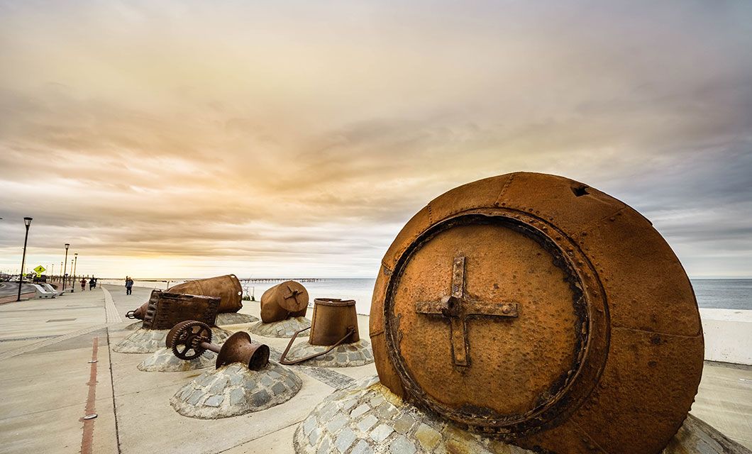 Large, rusted, circular metal structures are displayed on a seaside promenade at sunset in Ushuaia, with the ocean and a pier in the background under a cloudy sky.