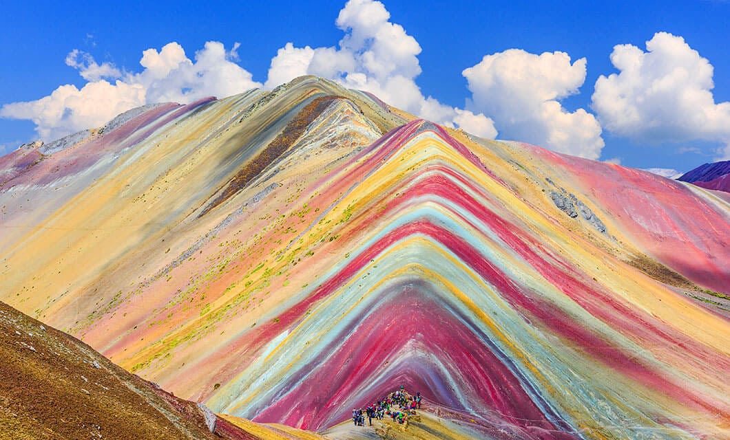 Colorful striped mountain with bands of red, yellow, pink, and blue under a bright blue sky; a group of people trekking at the base highlights its scale—a breathtaking South America landscape.