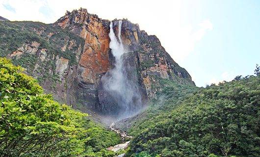 A tall waterfall cascades down a steep, rocky cliff on the Mount Roraima Trek, surrounded by lush green vegetation and trees, set against a partly cloudy sky.