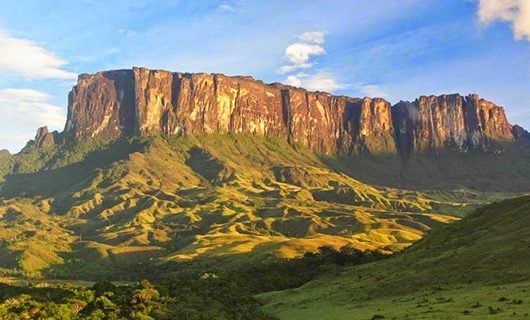 A massive tabletop mountain, Mount Roraima, with steep cliffs rises above green, rolling hills and valleys under a partly cloudy sky, bathed in warm sunlight.
