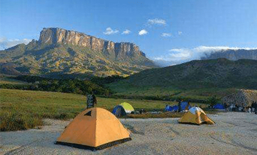 Tents are set up on a grassy plain with dramatic Mount Roraima rising in the background under a blue sky with clouds, suggesting a scenic, remote campsite along the Mount Roraima Trek.