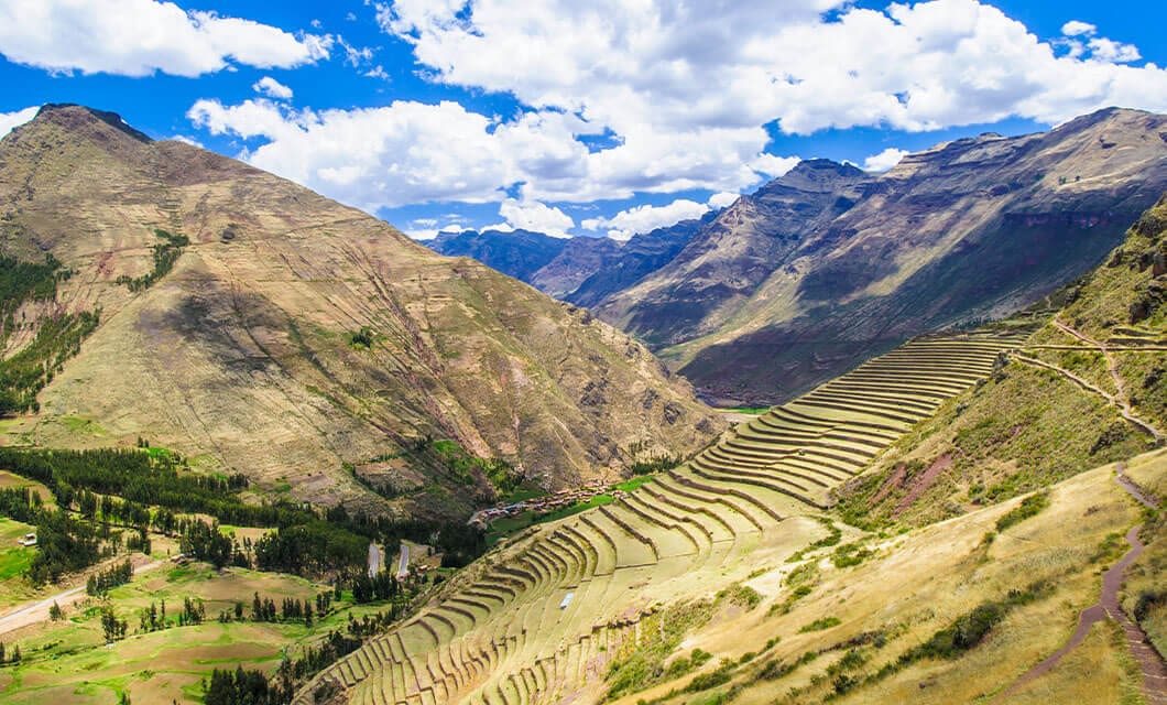 Terraced agricultural fields line the side of a steep, green mountainside under a blue sky with white clouds, in the heart of the Sacred Valley, with dramatic mountain peaks in the background.