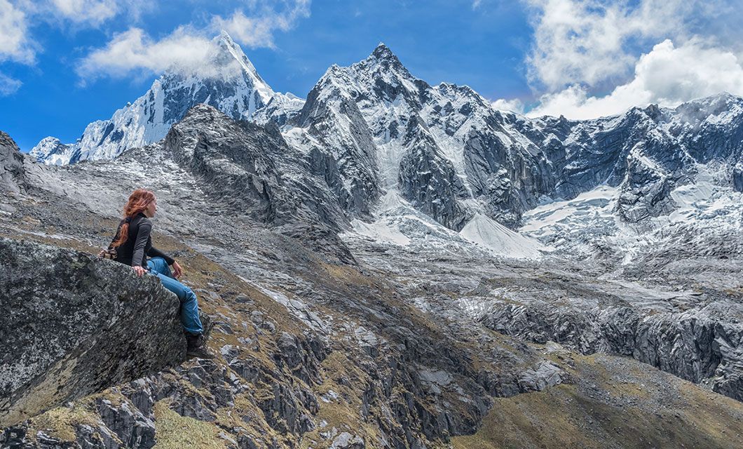 A person with long hair sits on a rock, gazing at dramatic, snow-capped mountains under a partly cloudy blue sky. Rugged terrain surrounds the figure, evoking the timeless spirit of the Incas in this vast, remote alpine landscape.