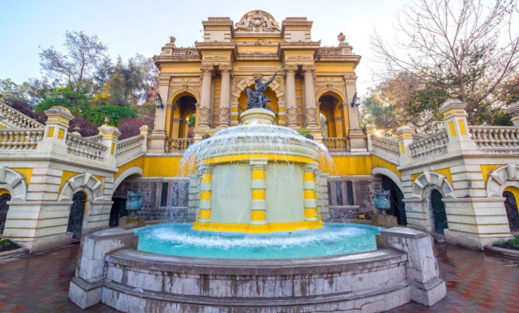 A large, ornate yellow and white fountain with water flowing from multiple tiers stands before a grand historic building in Ushuaia, framed by trees and decorative railings—a picturesque stop before your Patagonia Cruise.