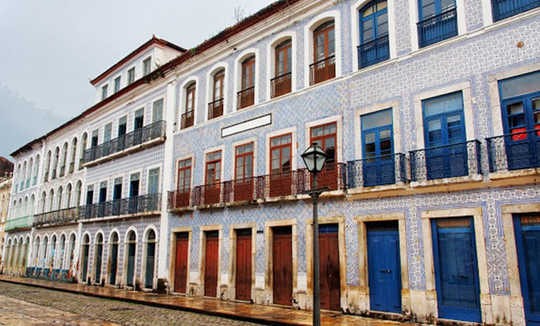 Rows of historic colonial buildings with blue-tiled facades, wooden doors, arched windows, and wrought-iron balconies line a cobblestone street under a cloudy sky—an inviting scene for a luxury adventure tour.