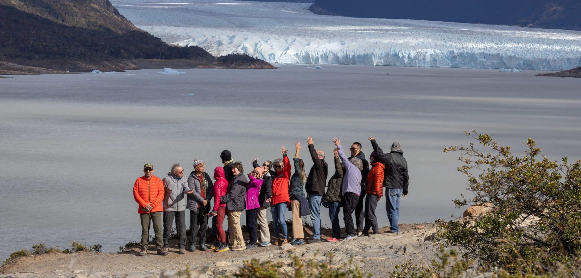A group of people in colorful jackets stand together near a lake in Patagonia, waving and facing a large glacier backed by mountains and sparse vegetation—capturing the spirit of a Guided Group Tour with Patagonia Essentials.