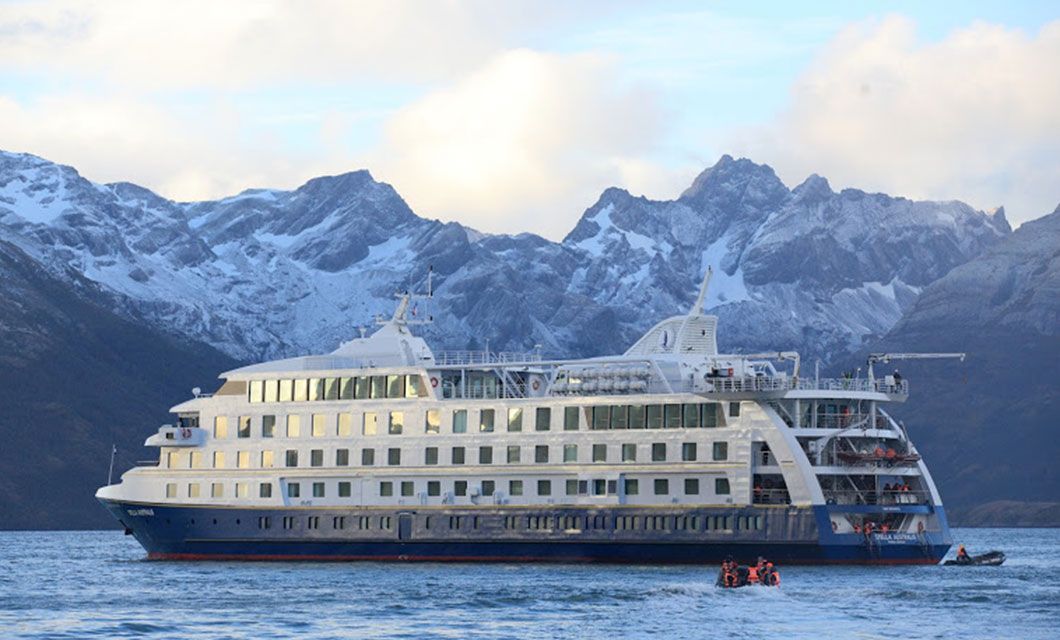 A large white and blue Patagonia Cruise ship sits on calm water with snow-capped mountains in the background; several small boats with people are near the ship, exploring the scenic beauty near Ushuaia.