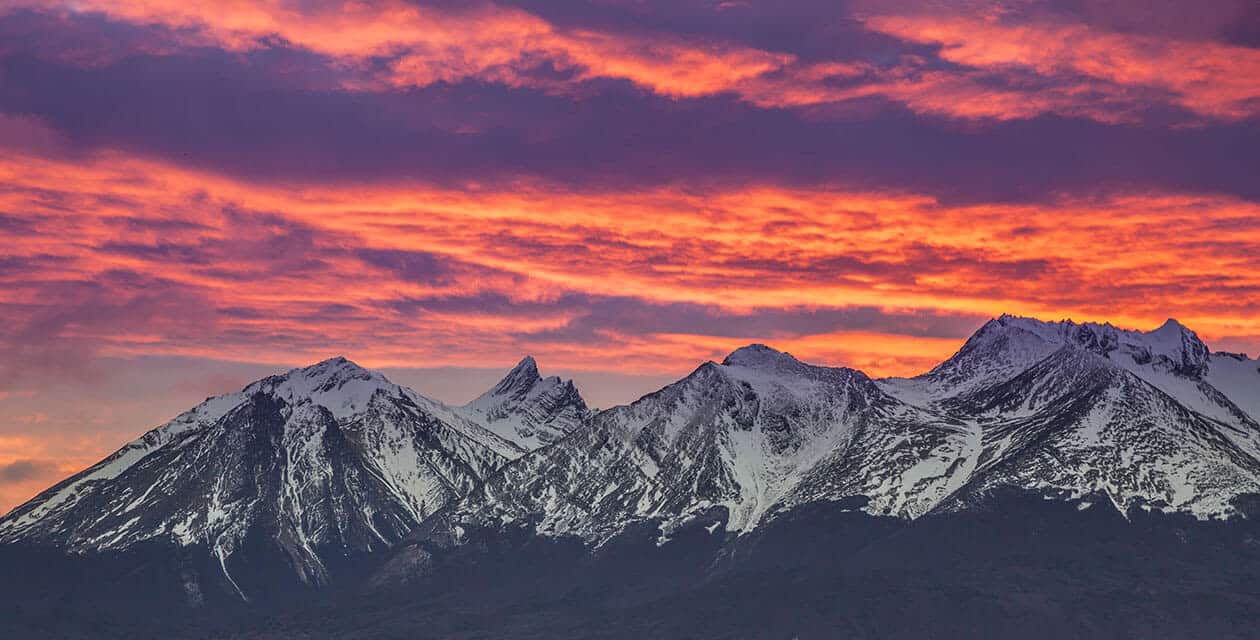 Snow-capped mountain peaks in Argentina under a dramatic sky at sunset, with vibrant orange, pink, and purple clouds—perfect for True Explorers seeking breathtaking views.