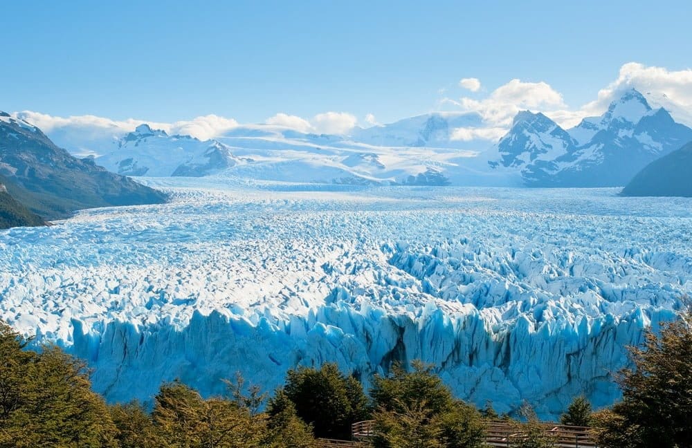 A vast glacier with jagged ice formations stretches across the landscape, surrounded by snow-capped mountains and green trees—a breathtaking sight for a memorable family journey under a bright blue sky with scattered clouds.