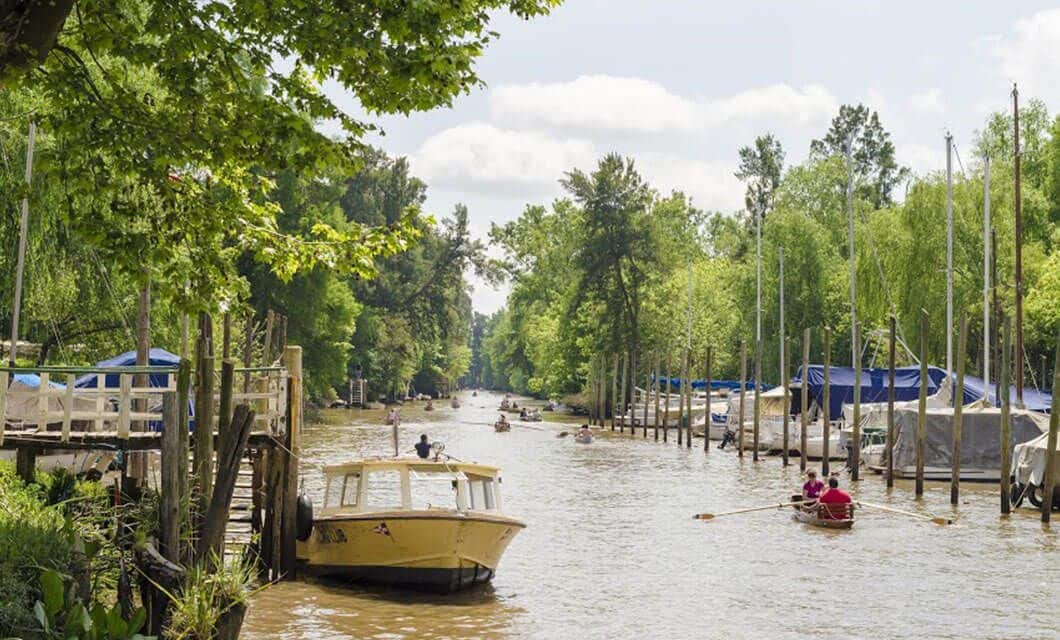 A river bordered by trees and docks, with boats docked along the shore and people rowing and riding boats on the water—perfect for a peaceful break during your coast to coast tour under a bright, partly cloudy sky.