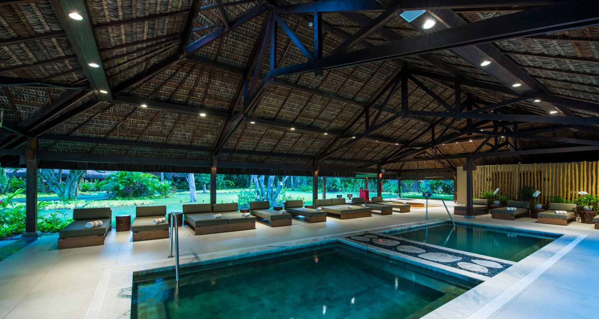 A spacious, covered lounge at Tivoli Eco-Resort features a thatched roof and rectangular indoor pool, with cushioned chairs and large windows overlooking Praia do Forte’s lush greenery. Soft lighting enhances the relaxing atmosphere.