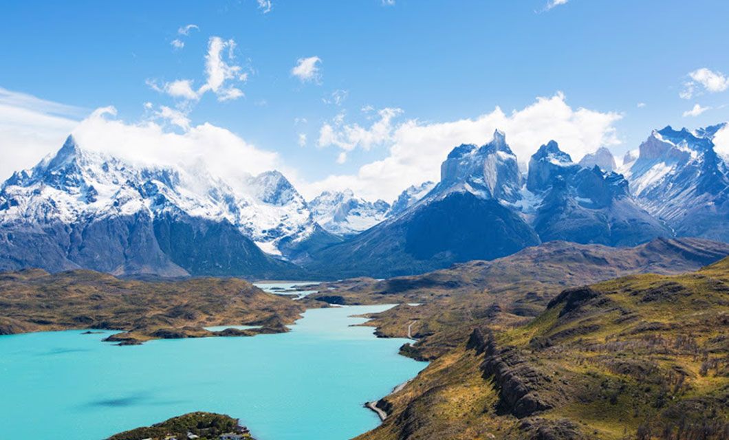 A turquoise lake winds through a valley surrounded by rugged, snow-capped mountains under a bright blue sky with scattered clouds, evoking the breathtaking landscapes of Torres del Paine. Rocky hills and sparse green vegetation cover the landscape.
