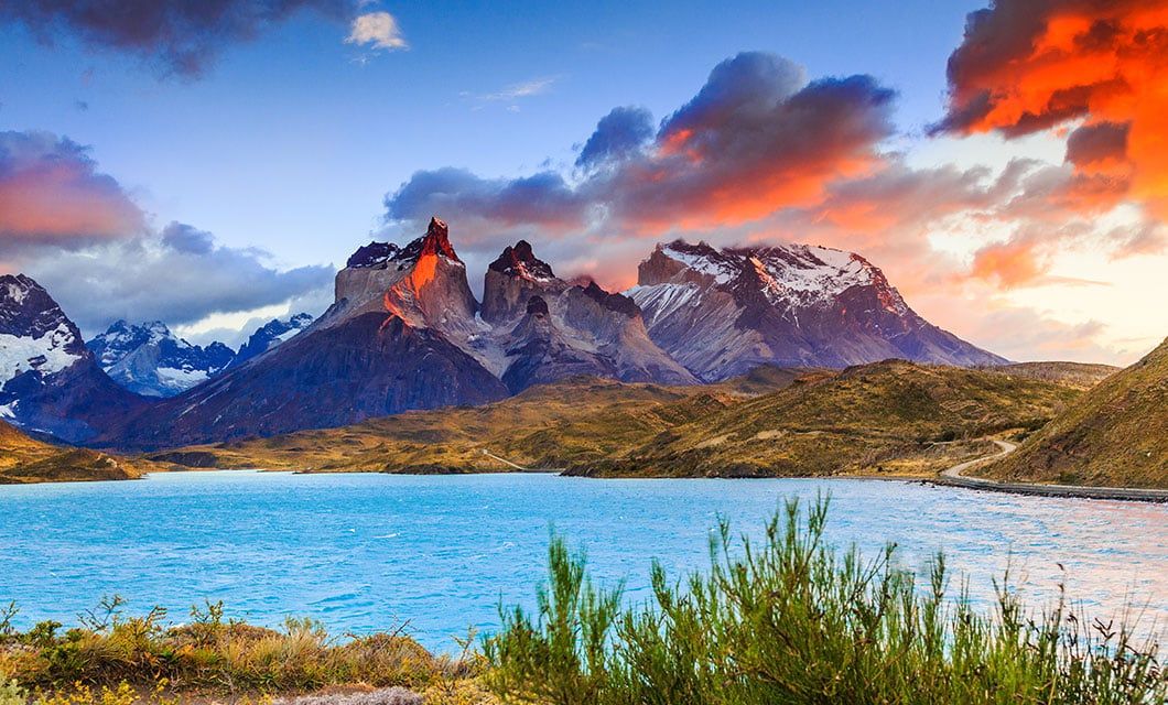 A stunning view of jagged mountains under a dramatic sunset sky with orange clouds, overlooking a bright blue lake surrounded by grassy hills—one of the true Highlights of Chile and a must-see for Chile tourism enthusiasts.