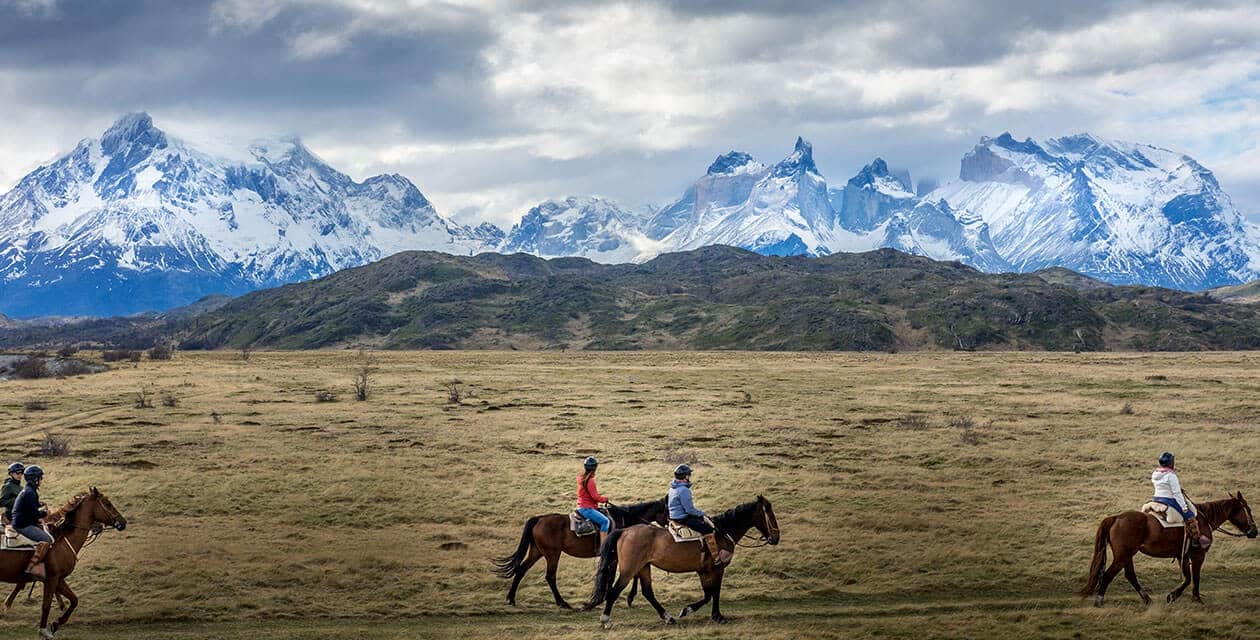 Four people ride horses across an open, grassy plain in Patagonia, with dramatic, snow-capped mountains and a cloudy sky in the background. The expansive landscape suggests a remote wilderness perfect for a guided group tour.