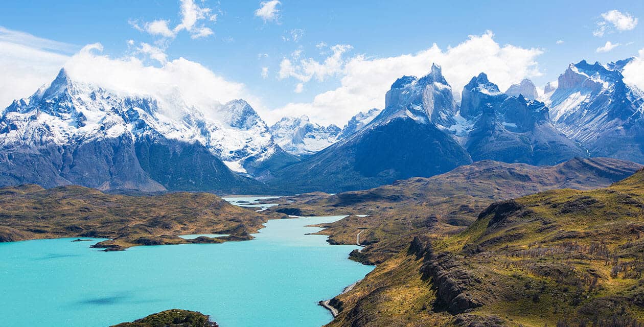 Turquoise lake surrounded by rolling hills and dramatic snow-capped mountains, perfect for trekking adventures in South America, under a bright blue sky with scattered clouds.