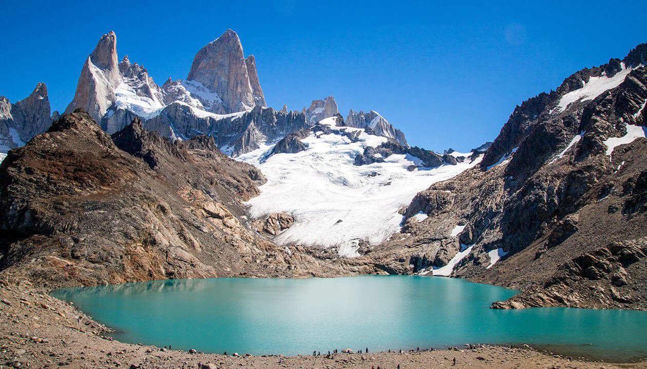 Turquoise mountain lake surrounded by rocky terrain and snow-capped peaks under a clear blue sky, with small figures of trekkers visible near the water’s edge—an iconic South America adventure scene.