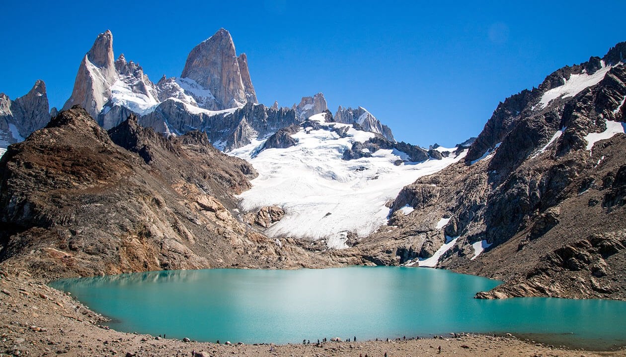 Turquoise glacial lake surrounded by rocky, snow-capped mountains under a clear blue sky, with waterfalls tumbling down jagged peaks and distant glaciers gleaming in the background.