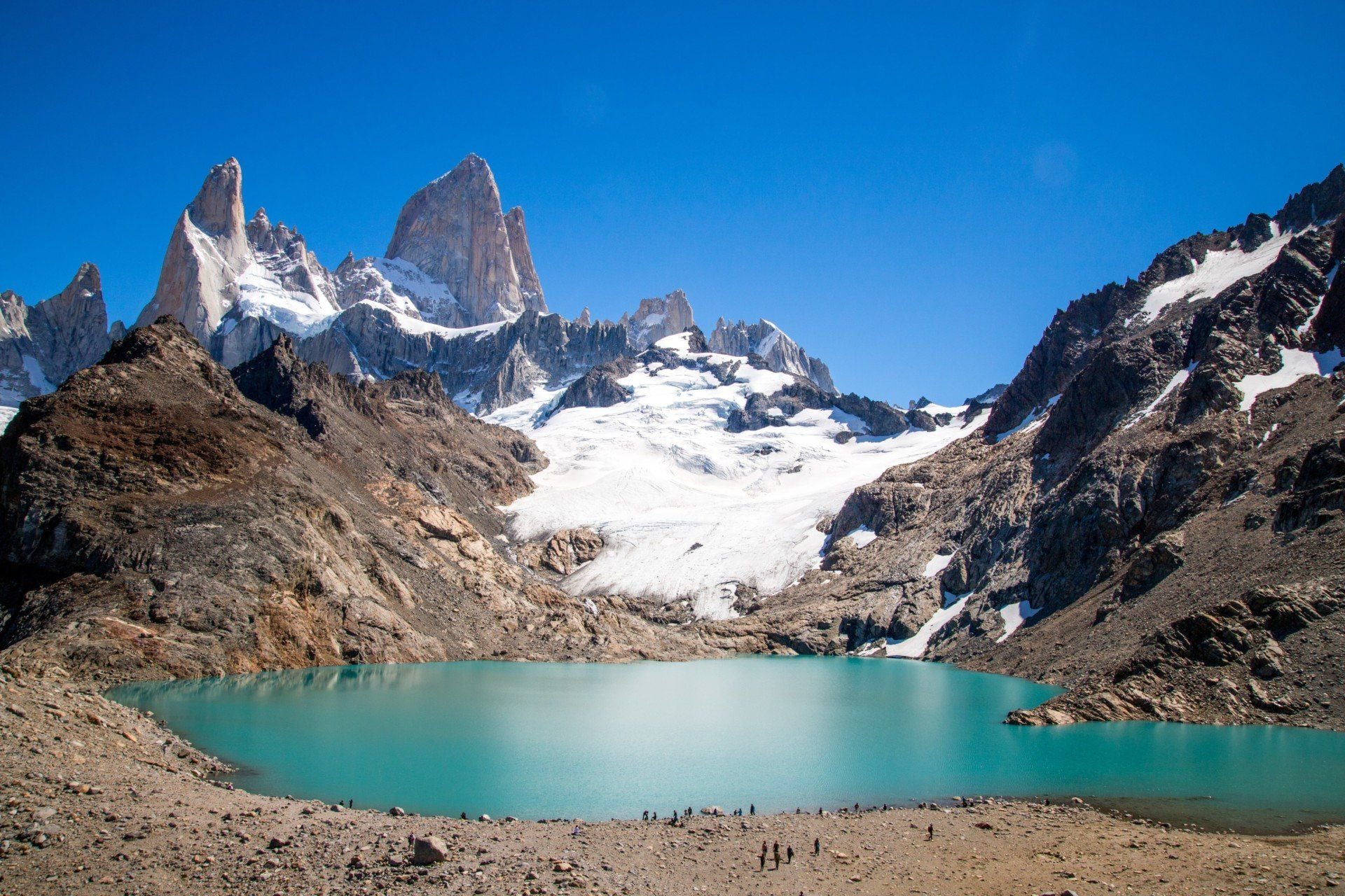 A turquoise glacial lake sits in front of rocky mountains with snowy peaks under a clear blue sky; small hikers are visible on the shore, embodying the spirit of true explorers traveling through Argentina’s breathtaking landscapes.