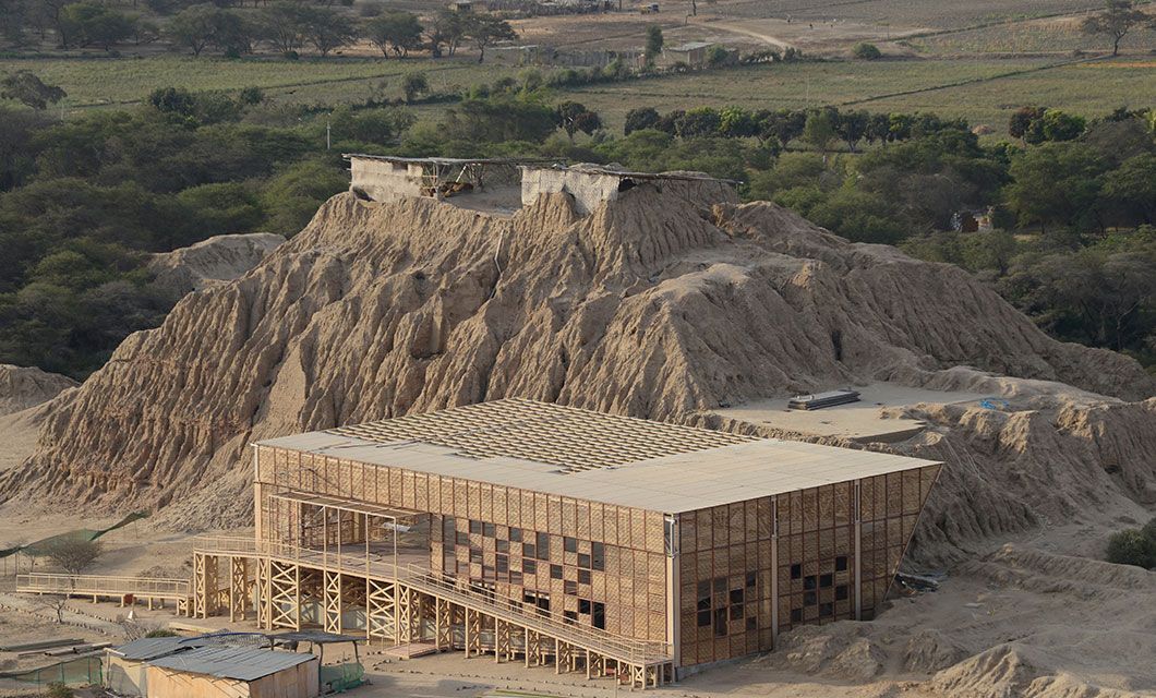 A large, modern building with a geometric patterned facade stands at the base of an eroded sandy hill near Trujillo, under a clear sky, with green trees and open fields stretching out in the background.