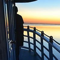 A person stands on the deck of a Tucano boat at sunset during a Brazil Amazon River Cruise, gazing over calm water as the colorful sky reflects shades of orange, yellow, and blue. The boat’s railing curves alongside the figure.