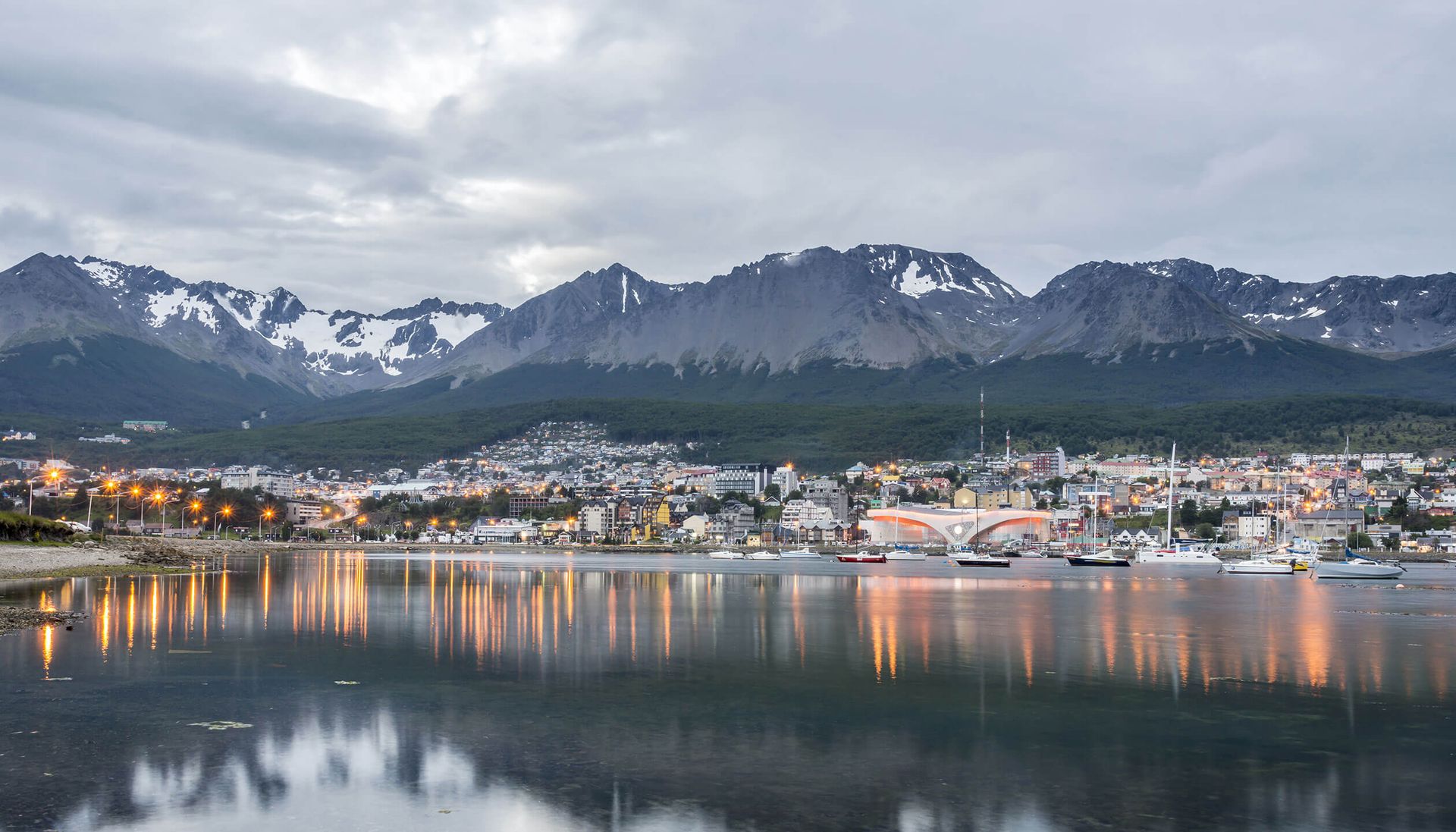 A coastal city with colorful buildings and boats sits by calm water, reflecting city lights and snow-capped mountains—a picturesque stop on any Patagonia tour under a cloudy sky.