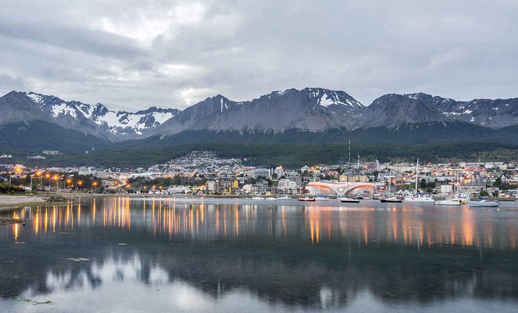 A scenic view of Wild Argentina’s coastal city with colorful buildings, calm water reflecting city lights, boats docked along the shore, and snow-capped mountains in the background under a cloudy sky.