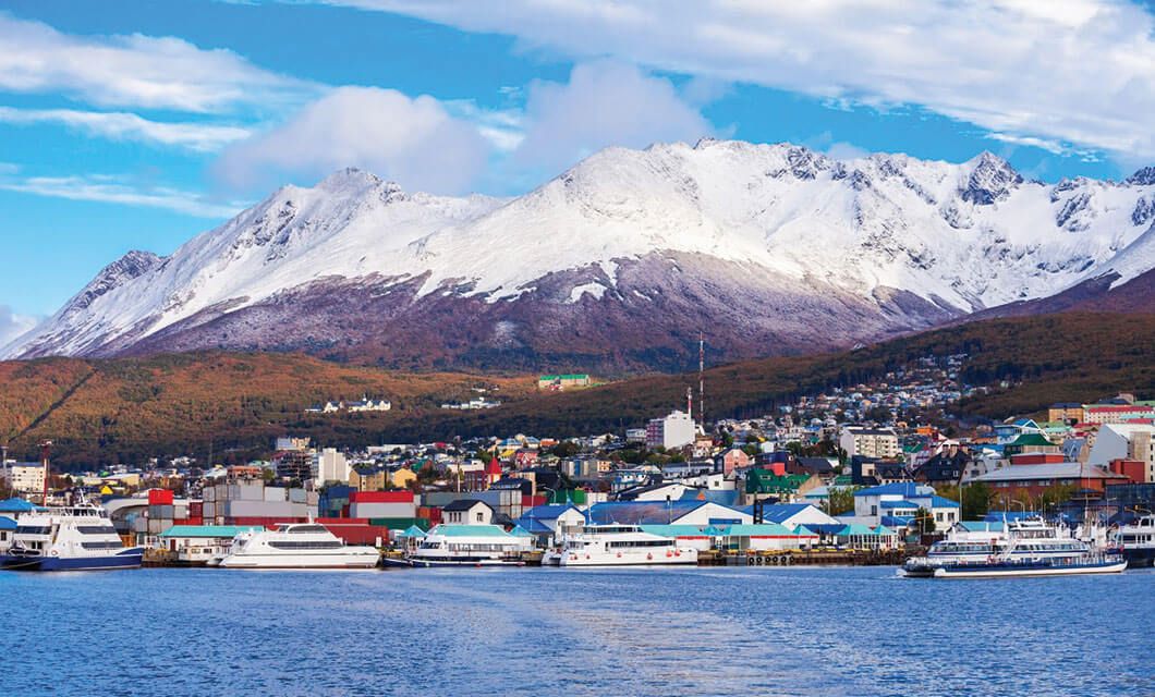 A coastal town with colorful buildings and docked boats sits at the base of snow-capped mountains under a blue sky—an inviting scene for any Patagonia travel adventure.