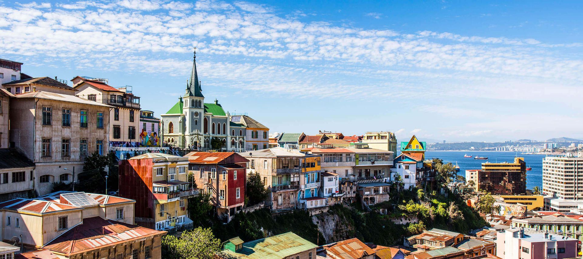Colorful hillside buildings and a church with a green spire overlook the ocean in Valparaíso, Chile—an inviting scene for those enjoying Valparaiso Tours under blue skies dotted with clouds, with ships visible in the distance.